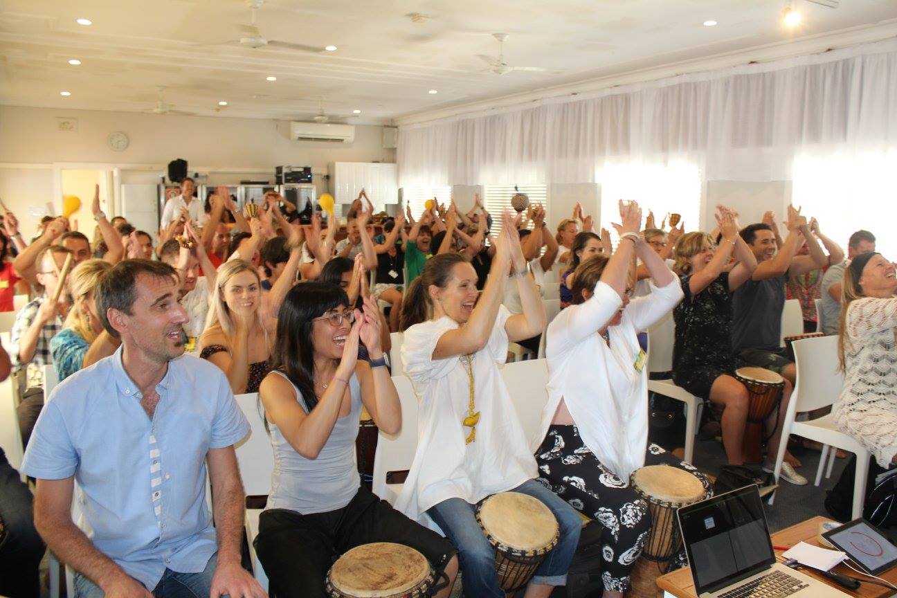 A group of people sitting in a room playing drums and clapping their hands, appearing engaged and enthusiastic. The room, similar to an ideal Meta Ads Audience Targeting setting, is brightly lit with white curtains and modern decor.