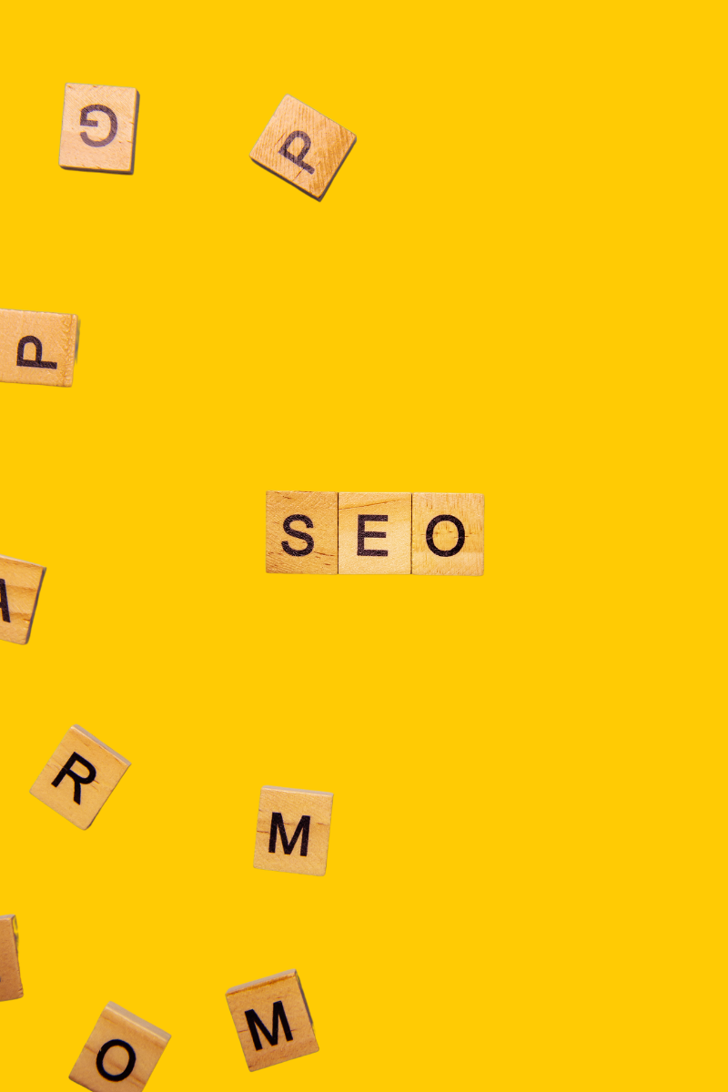 Wooden letter tiles neatly spell out SEO on a vibrant yellow background, surrounded by a scattering of other tiles.