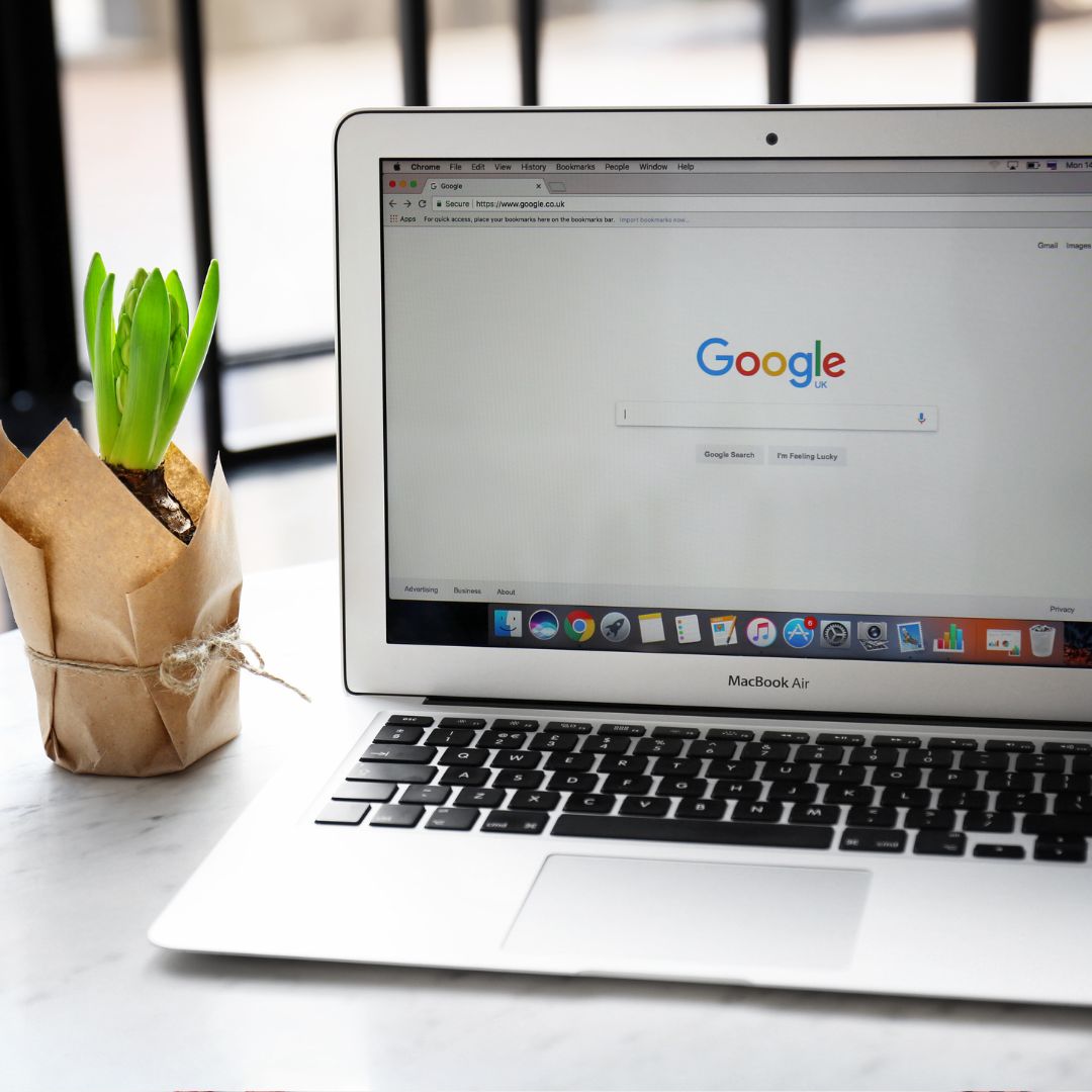 Open laptop displaying Google homepage on a table with a small plant in a brown paper pot.